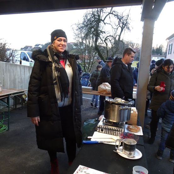 Eine Frau steht neben einem Tisch mit Essen. Sie trägt einen Mantel und eine Schärpe. Menschen versammeln sich um einen Tisch mit einem Topf. Ein Mann steht neben ihr. Bäume und ein Gebäude sind im Hintergrund.