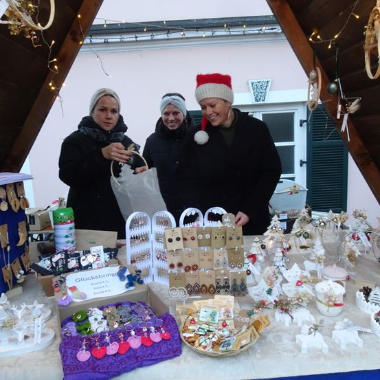 Drei Frauen stehen hinter einem Tisch voller Weihnachtsdekorationen. Sie lächeln und scheinen dabei zu sein, die Ausstellung aufzubauen. Der Tisch ist voll mit verschiedenen Ornamenten, und über ihnen hängen Lichter und Dekorationen.