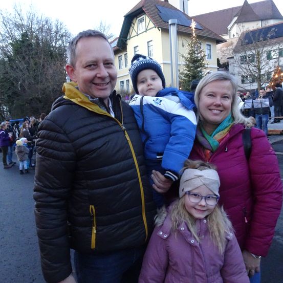 Eine vierköpfige Familie steht im Freien in Winterkleidung. Der Mann trägt eine schwarze und gelbe Jacke. Die Frau trägt eine lila Jacke. Der Säugling wird vom Mann gehalten und trägt eine blaue Jacke. Das Mädchen trägt eine lila Jacke und eine Kopfband.