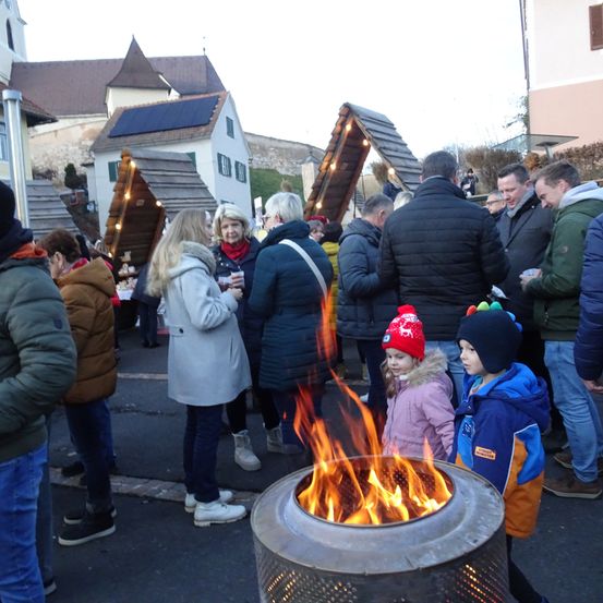 Mehrere Menschen versammeln sich um einen Feuerkorb im Freien. Eine Frau in einem weißen Mantel steht mit einem Becher, während zwei Kinder in Winterkleidung dicht am Feuer stehen. Im Hintergrund befinden sich Gebäude und Straßenlaternen.