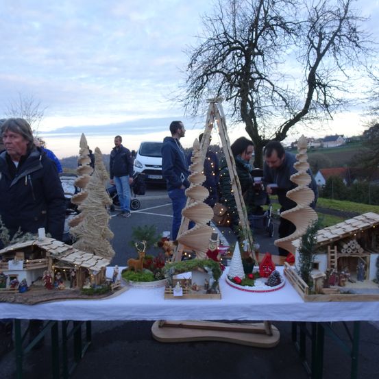 Ein Tisch mit verschiedenen Weihnachtsdekorationen im Freien. Menschen sind versammelt und bewundern die Miniaturdorfszenen. Bäume und kleine Figuren sind Teil der Ausstellung.