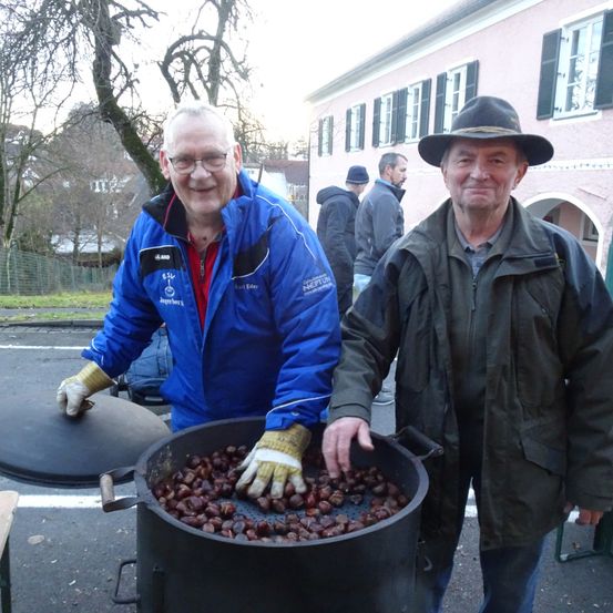 Zwei ältere Männer in Winterkleidung lächeln vor einem großen Topf mit gerösteten Kastanien, mit einem Gebäude und Menschen im Hintergrund.