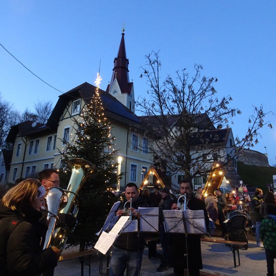 Eine Band spielt draußen vor einem Gebäude bei Nacht. Die Band besteht aus einem Trompeter, einem Tubaspieler und einem Posaunisten. Das Gebäude hat einen Glockenturm und einen Weihnachtsbaum vor sich.