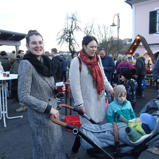 Zwei Frauen stehen draußen, eine hält einen Becher und die andere schiebt einen Kinderwagen mit zwei Kindern. Hinter ihnen steht eine Menschenmenge, einige tragen Mützen und Schals. Es gibt auch Tische, einen Laternenpfahl und ein Haus im Hintergrund.