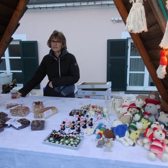 Eine Frau steht hinter einem mit einem weißen Tuch bedeckten Tisch, auf dem verschiedene Backwaren und handgefertigte Spielzeuge ausgestellt sind, mit einem Haus und Dekorationen im Hintergrund.
