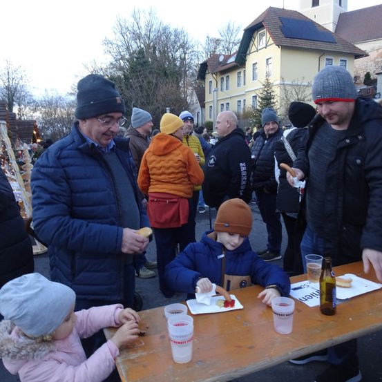 Eine Gruppe von Menschen, darunter Erwachsene und Kinder, versammelt sich um einen Tisch und isst. Sie sind warm in Winterkleidung gekleidet. Im Hintergrund befinden sich Gebäude, eines mit Solarpanelen.