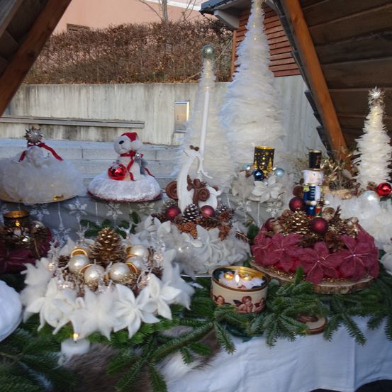 Ein festlicher Tisch mit mehreren Weihnachtsdekorationen, darunter ein Schneemann, zwei Weihnachtsbäume, Ornamente, Tannenzapfen und Kerzen. Der Tisch ist mit einem weißen Tuch bedeckt und von Grünpflanzen umgeben.