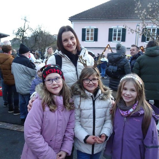 Eine Frau mit drei jungen Mädchen steht vor einem Gebäude bei einem festlichen Anlass. Im Hintergrund sind Menschen versammelt.