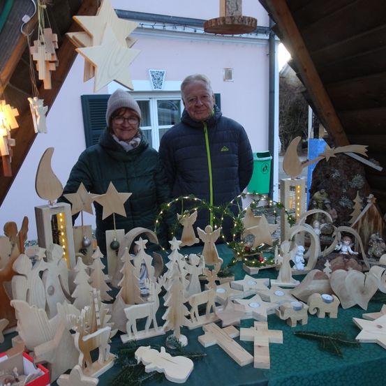 Zwei Personen stehen hinter einem Tisch mit hölzernen Weihnachtsdekorationen. Eine Person trägt eine Brille und einen Hut, die andere eine Jacke. Über ihnen hängen Sterne und eine Girlande.