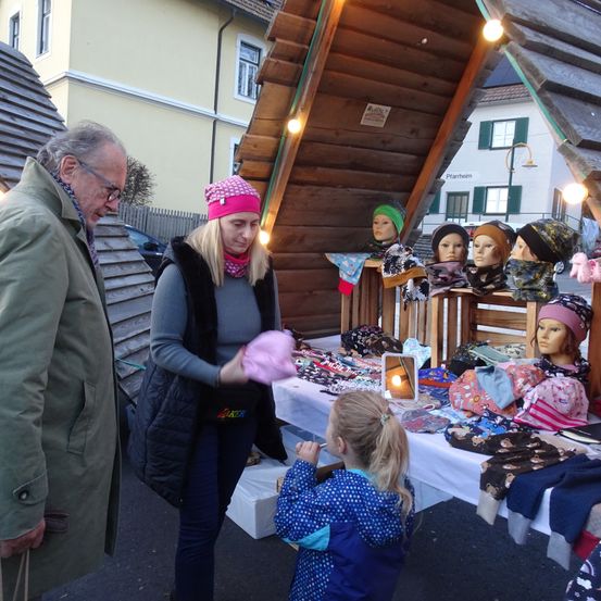 Eine Frau mit einer rosa Mütze steht hinter einem Tisch voller Hüte. Ein Mann und ein junges Mädchen stehen vor dem Tisch. Auf dem Tisch sind maniquins mit Hüten ausgestellt.