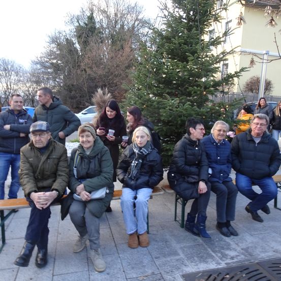 Eine Gruppe von Menschen sitzt auf einer Bank vor einem Weihnachtsbaum, alle warm gekleidet. Einige Personen halten Tassen und scheinen zu unterhalten. Der Hintergrund zeigt ein gelbes Gebäude und geparkte Autos.