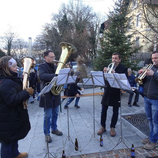 Eine Gruppe von Musikern spielt auf der Straße und musiziert Trompeten und Tuben. Sie stehen auf einer Bühne mit Notenständern. Dahinter sitzt eine Person auf einer Bank. Im Hintergrund befinden sich Bäume und ein Gebäude.