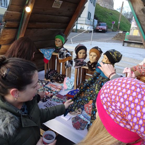 Mehrere Frauen schauen sich verschiedene farbenfrohe Accessoires an, die auf einem Tisch bei einer Outdoor-Veranstaltung ausgestellt sind, einschließlich Hüte und Schals an Mannequins.