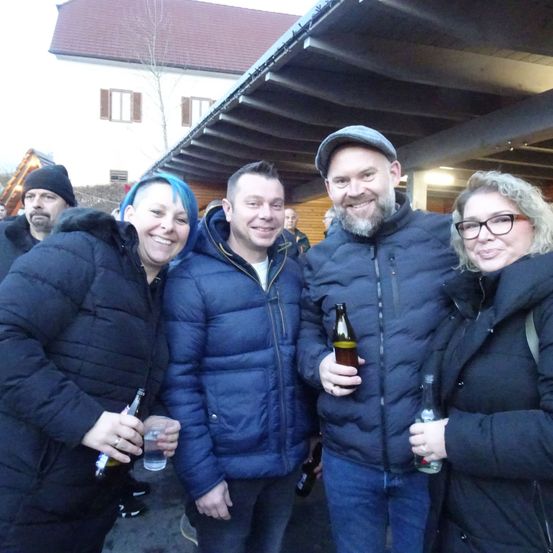 Fünf Menschen lächeln für ein Foto und halten Bierflaschen. Der Mann rechts trägt einen Hut und einen Bart. Hinter ihnen steht ein Gebäude mit einem roten Dach.