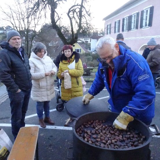 Eine Gruppe von Menschen in Winterkleidung versammelt sich um einen großen Topf voller Kastanien, wobei ein Mann Handschuhe trägt und sie knackt. Ein Kinderwagen ist in der Nähe geparkt, und ein Gebäude mit mehreren Fenstern ist im Hintergrund zu sehen.