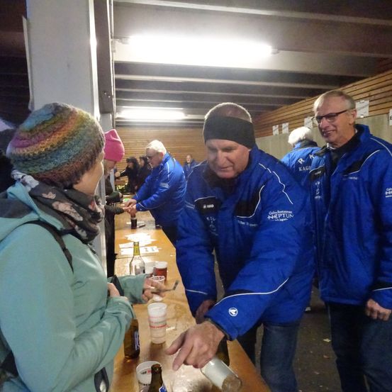 Eine Gruppe von Menschen in blauen Jacken versammelt sich um einen Tisch. Zwei Männer schenken Bier in Gläser für eine Frau ein.