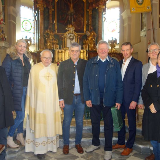 Eine Gruppe von Menschen steht vor einem Altar in einer Kirche, wobei ein Priester in der Mitte eine gelbe Robe trägt. Hinter ihm stehen mehrere Männer und Frauen in formeller Kleidung. Im Hintergrund befinden sich eine goldene Statue und Buntglasfenster.