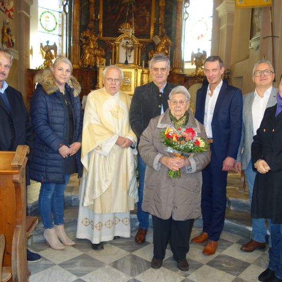 Eine Gruppe von Menschen, darunter eine ältere Frau mit Blumen, steht in einer Kirche vor einem Altar mit einem Priester.