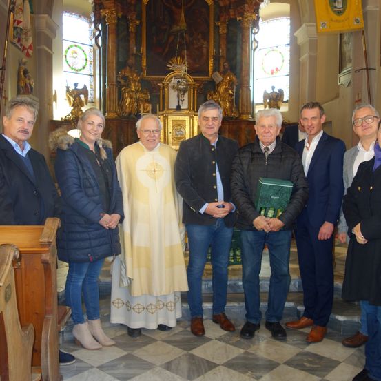 Eine Gruppe von Menschen steht in einer Kirche mit einem Priester. Sie sind in Winterkleidung gekleidet und scheinen für ein Foto zu posieren. Hinter ihnen steht ein Altar mit einem Gemälde und einer Statue.