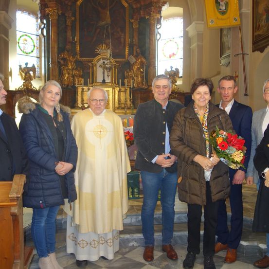 Eine Gruppe von Menschen steht in einer Kirche, mit einem Priester in der Mitte, der eine weiße Robe und eine Stola trägt. Hinter ihm befindet sich ein goldener Altar.
