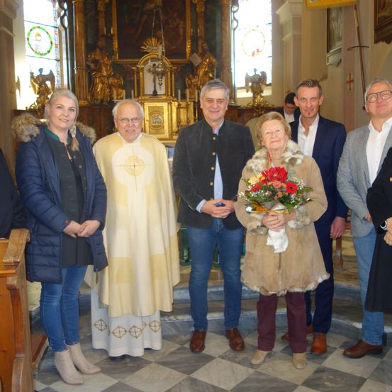 Mehrere Personen stehen in einer Kirche, eine davon hält Blumen. Der Mann in der Mitte trägt eine Soutane, während andere ein Geschäft tragen. Hinter ihnen befindet sich ein Altar mit religiösen Artefakten.