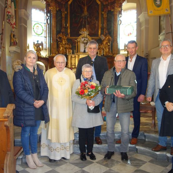 Eine Gruppe von Menschen steht in einer Kirche. Eine Frau hält Blumen, ein Mann in einem gelben Gewand steht neben ihr. Ein Mann vor ihnen hält eine grüne Schachtel. Hinter ihnen ein Priester und drei Männer in Anzügen. Altar im Hintergrund.