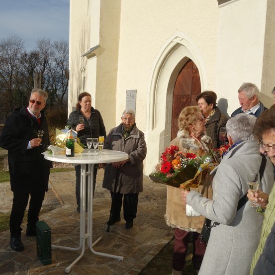 Eine Gruppe von Menschen in Winterkleidung steht vor einer Kirche und feiert mit Weinglasern und Blumen. Ein Tisch hält eine Champagnerflasche.