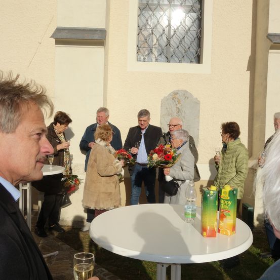 Eine Gruppe von Menschen steht draußen, einige halten Weingläser und Blumen. Ein Tisch mit Getränken steht davor. Die Umgebung scheint ein gesellschaftliches Treffen vor einem Gebäude zu sein.