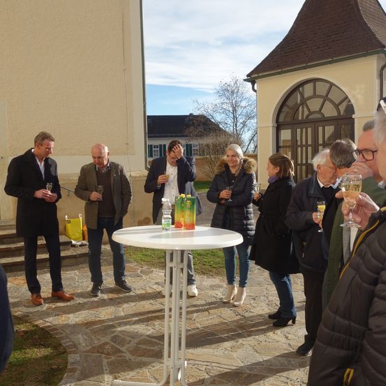 Eine Gruppe von Menschen steht um einen Tisch in einem Innenhof und hält Weingläser in der Hand. Die Umgebung umfasst ein Gebäude mit einem braunen Dach und Glasfenstern. Einige Menschen tragen Mäntel, während andere in Freizeitkleidung sind. Flaschen und Dosen stehen auf dem Tisch.