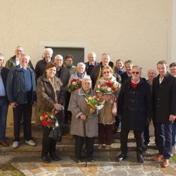 Eine Gruppe von Menschen in Winterkleidung steht auf Stufen vor einem Gebäude. Einige halten Blumen. Die Stimmung ist fröhlich.