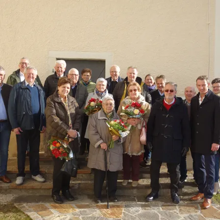 Eine Gruppe von Menschen in Winterkleidung steht auf Stufen vor einem Gebäude. Einige halten Blumen. Die Stimmung ist fröhlich.