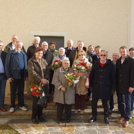 Eine Gruppe von Menschen in Winterkleidung steht auf Stufen vor einem Gebäude. Einige halten Blumen. Die Stimmung ist fröhlich.