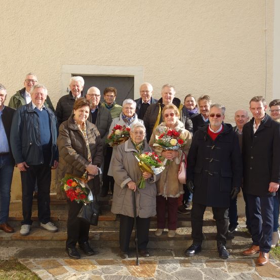Eine Gruppe von Menschen in Winterkleidung steht auf Stufen vor einem Gebäude. Einige halten Blumen. Die Stimmung ist fröhlich.