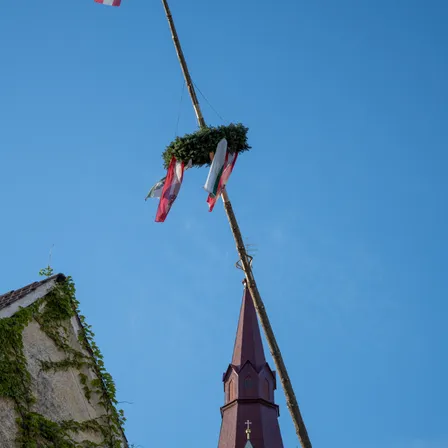 Ein Kirchturm mit einem Kreuz oben und einem Baum mit einem Kranz und Flaggen, die daran befestigt sind, gegen einen klaren blauen Himmel.
