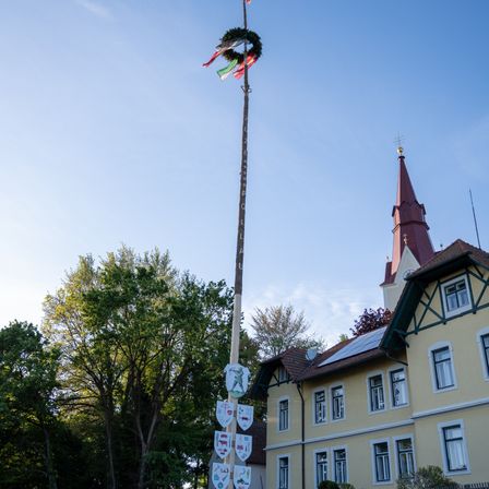 Ein hoher Pfahl mit mehreren Flaggen und Schilden steht vor einem gelben Gebäude mit einem Turm unter einem klaren blauen Himmel.