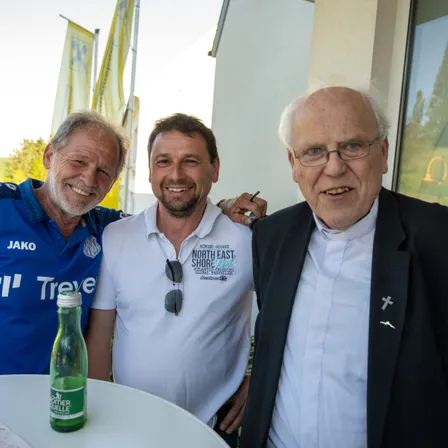 Drei Männer lächeln und stehen zusammen. Einer in einem blauen Shirt, ein anderer in einem weißen Shirt und ein dritter in einem schwarzen Anzug. Eine Flasche steht auf einem Tisch vor ihnen.