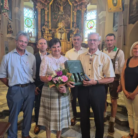 Eine Gruppe von Menschen steht lächelnd in einer Kirche. Eine Frau hält Blumen und eine Geschenkbox, während ein Mann neben ihr ein Geschenk hält. Hinter ihnen hält ein anderer Mann ein Glas. Im Hintergrund befinden sich Buntglasfenster und ein goldener Altar.