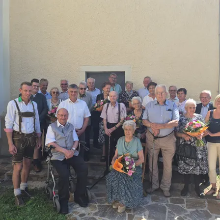 Eine große Gruppe älterer Menschen ist draußen versammelt, einige in traditioneller Kleidung, und posiert für ein Foto vor einem Gebäude. Viele halten Blumen.