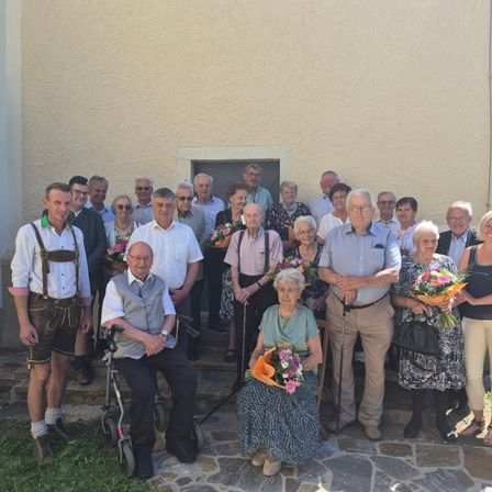Eine große Gruppe älterer Menschen ist draußen versammelt, einige in traditioneller Kleidung, und posiert für ein Foto vor einem Gebäude. Viele halten Blumen.