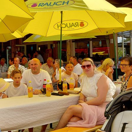 Eine Gruppe von Menschen sitzt unter gelben Sonnenschirmen an einem Tisch, einige mit Sonnenbrillen und andere mit Getränken vor sich.