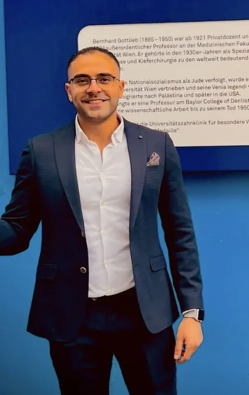 Ein Mann in einem schwarzen Anzug, einem weißen Hemd und einer Brille steht vor einer blauen Wand und posiert wahrscheinlich für ein Foto.