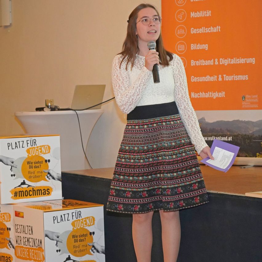 Eine Frau steht auf der Bühne, hält ein Mikrofon und spricht. Sie trägt eine Brille und einen Blumenrock. Hinter ihr stehen zwei Boxen mit dem Text 'Platz fur Jugend' und ein Banner mit verschiedenen Symbolen.