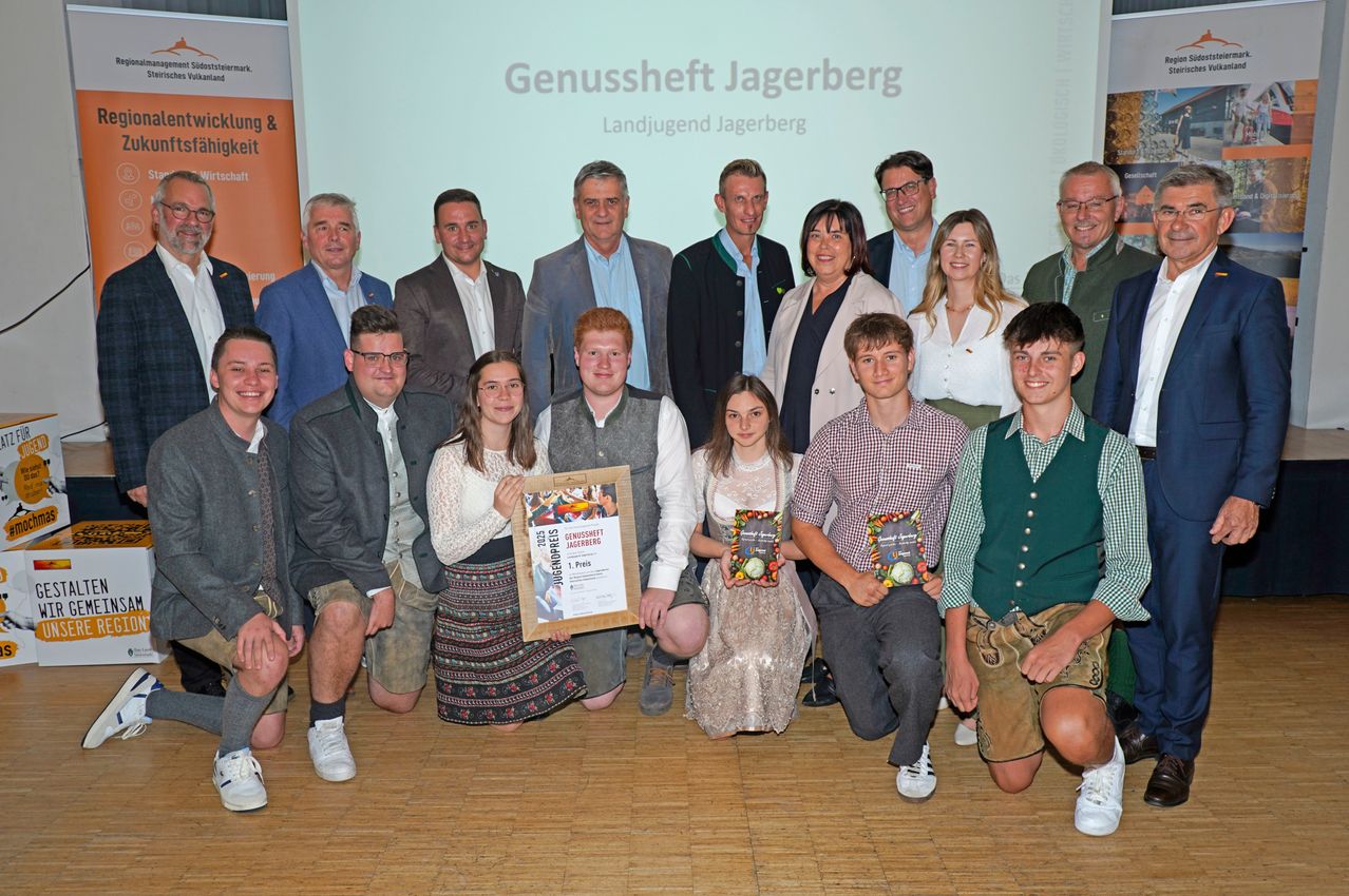 Eine Gruppe von Menschen posiert für ein Foto, mehrere halten Auszeichnungen und Urkunden. Die Umgebung scheint eine Veranstaltung oder Preisverleihung zu sein, mit einem Hintergrund, auf dem 'Genussmarkt Jagerberg' steht.