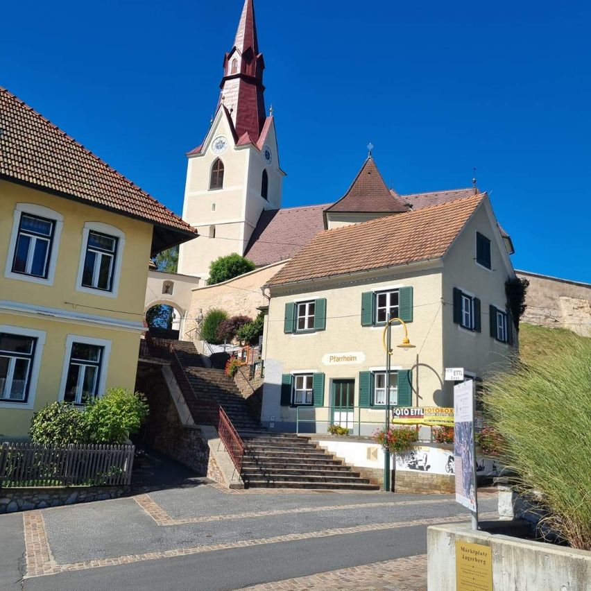 Ein Straßenblick auf eine Kirche mit Turm, rotem Dach und großen Fenstern. Die Kirche ist von Häusern, Pflanzen und einem kleinen Garten umgeben.