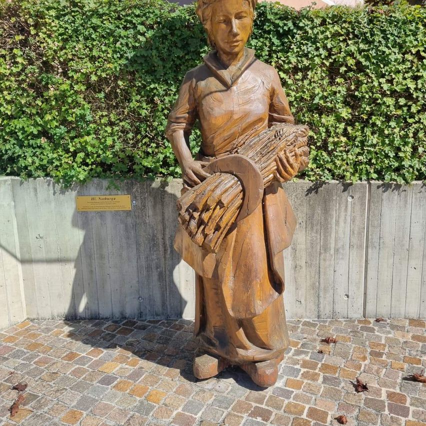 Eine Holzstatue einer Frau, die einen Weizenstapel hält, steht auf einem Ziegelboden mit einer Betonmauer dahinter.