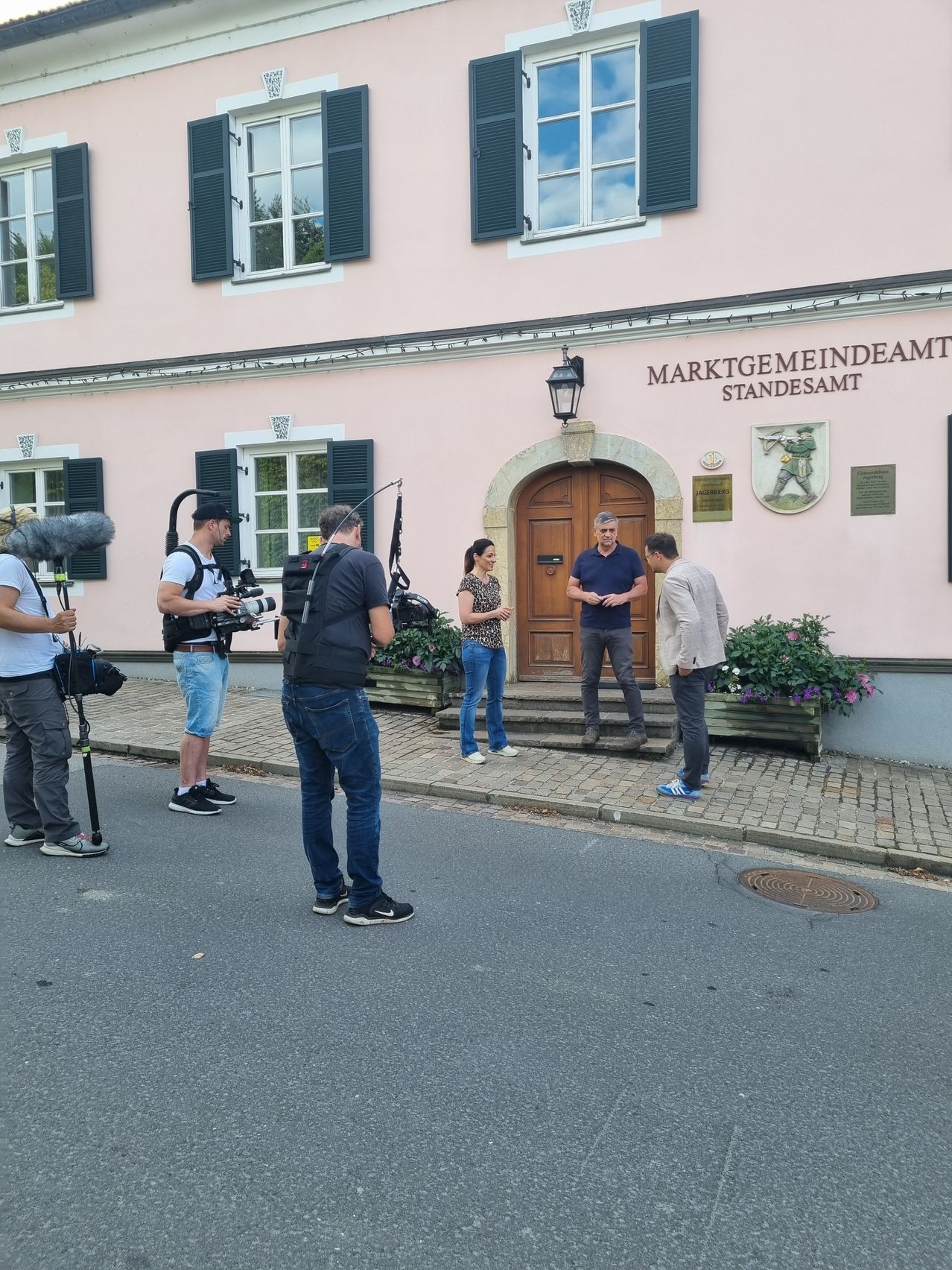 Eine Gruppe von Leuten filmt vor einem rosafarbenen Gebäude mit einer Holztür. Ein Mann hält ein Mikrofon, und ein anderer hat eine Kamera auf einem Stativ. Zwei Frauen stehen vor der Tür, und ein Mann steht auf den Stufen.