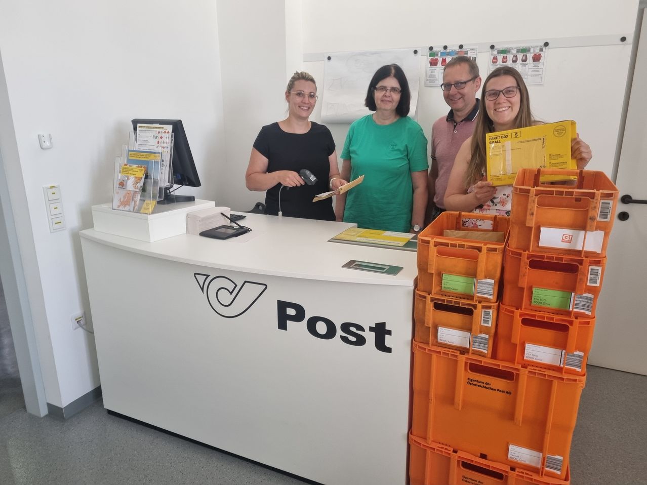 Vier Personen stehen lächelnd hinter einem Tresen in einem Postamt und halten Dokumente in der Hand. Der Tresen hat eine weiße Oberfläche mit dem Wort Post. Mehrere Gegenstände sind auf dem Tresen.