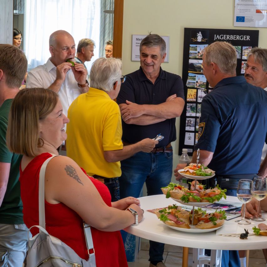 Eine Gruppe von Menschen steht um einen Tisch mit Essen. Ein Mann isst ein Sandwich, ein anderer Mann hält ein Handy. Hinter ihnen hält ein Mann ein Glas Wein.