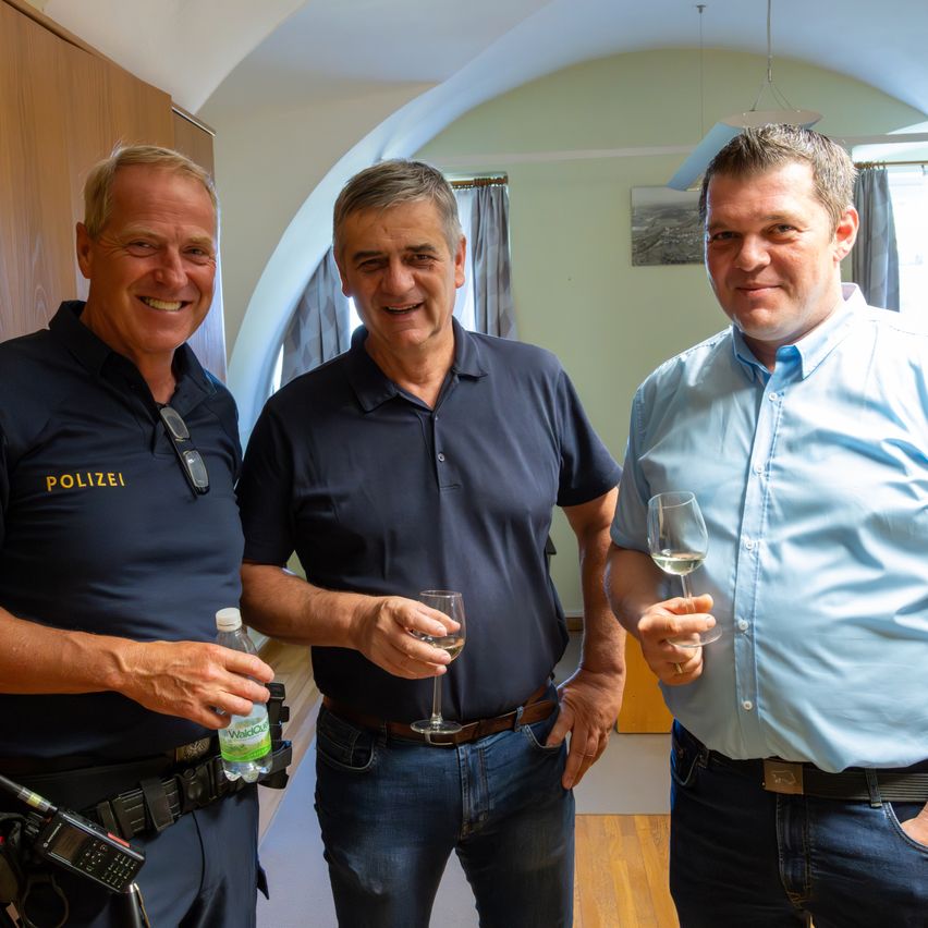 Drei Männer stehen lächelnd in einem Raum zusammen. Der Mann auf der linken Seite hält eine Flasche Wasser. Der Mann in der Mitte hält ein Weinglas, und der Mann auf der rechten Seite hält ebenfalls ein Weinglas.