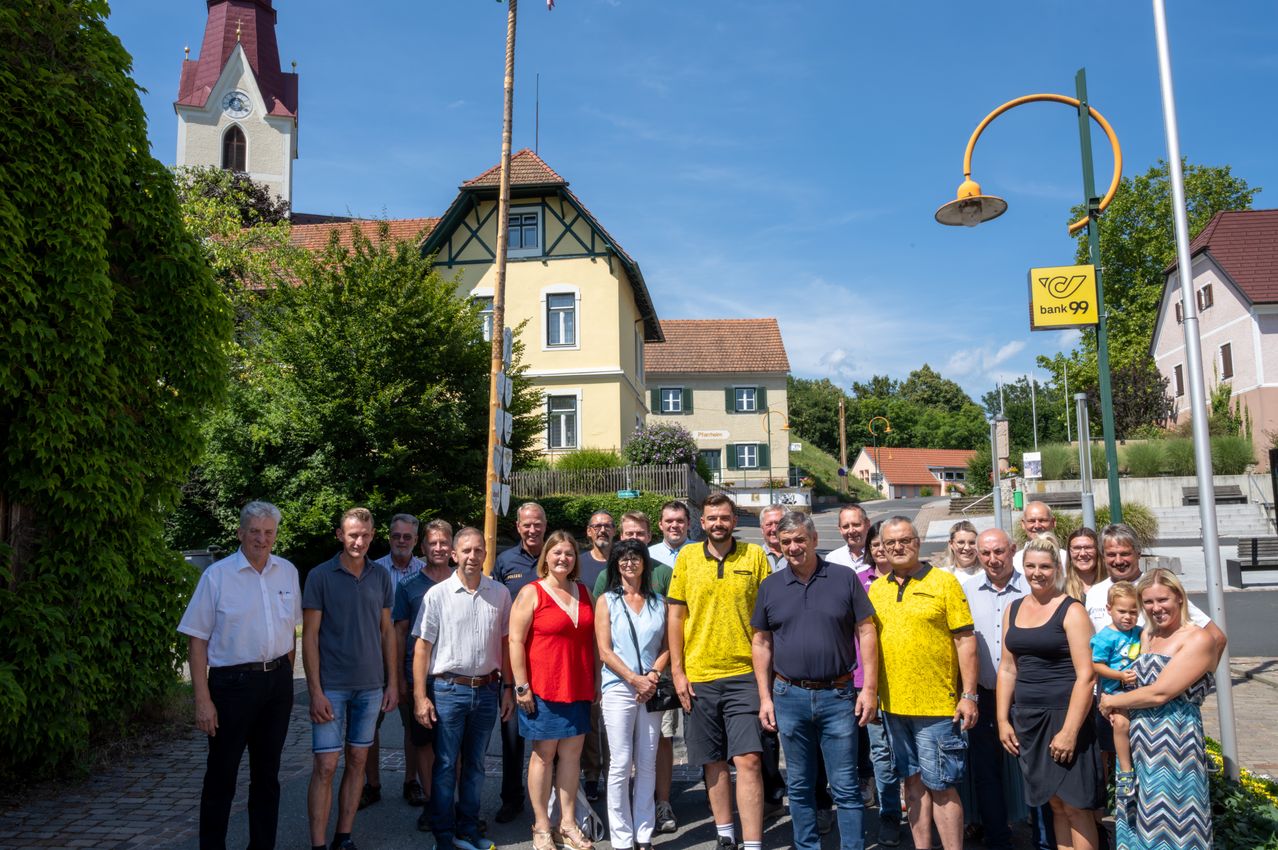 Eine Gruppe von Menschen steht vor einem Haus mit Fahnenmast an einem sonnigen Tag.